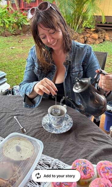 A woman in a denim jacket smiles while pouring tea into a teacup outdoors. Colorful cupcakes and dessert containers are on the table, creating a cozy scene.