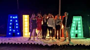 Six people pose at night in front of a large illuminated "ALOHA" sign, each letter glowing in different colors: blue, yellow, orange, and green.