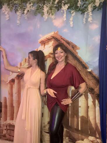 Two women in Greek-inspired costumes pose in front of a backdrop depicting ancient ruins. One wears a cream dress, the other wears burgundy. Decorative foliage hangs above.