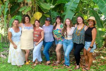 Eight women stand together outdoors in front of large green plants, smiling and casually dressed in summery clothes. The lush setting suggests a tropical, relaxed atmosphere.