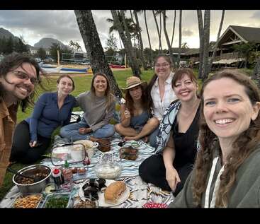 Seven smiling friends sit on a picnic blanket outdoors, surrounded by delicious food and trees, with a scenic backdrop of mountains, palm trees, and rustic buildings.