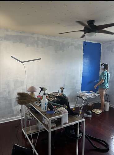 A person is painting a room blue. The walls are mostly primed, furniture is piled in the center, and painting supplies are scattered. Ceiling fan above.