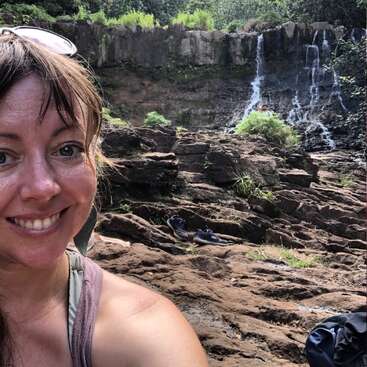 The image depicts a woman taking a selfie in front of a rocky waterfall, surrounded by lush greenery and trees, with a backpack visible on the ground.