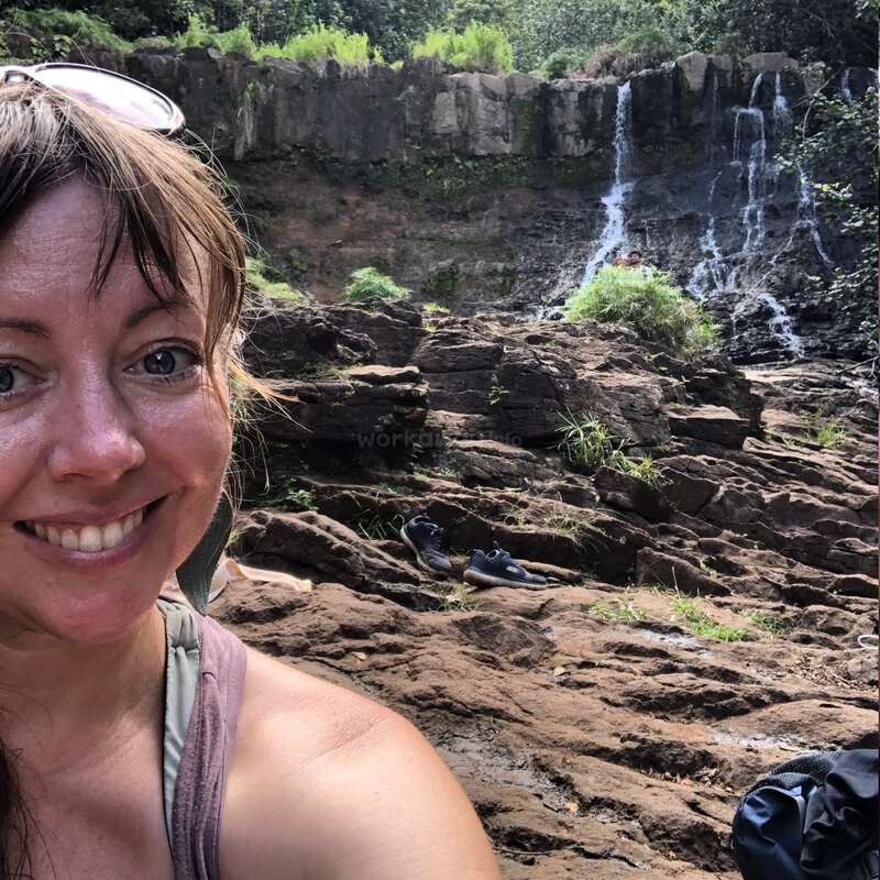The image depicts a woman taking a selfie in front of a rocky waterfall, surrounded by lush greenery and trees, with a backpack visible on the ground.