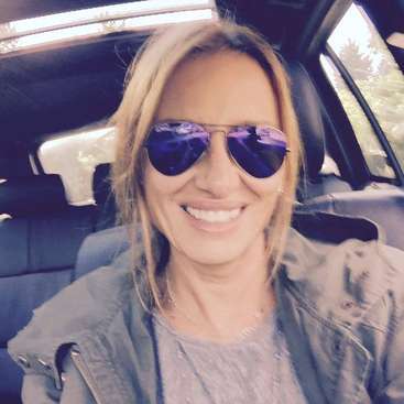 La imagen muestra a una mujer rubia con gafas de sol, sentada en el asiento trasero de un coche, con árboles visibles a través de las ventanillas detrás de ella.