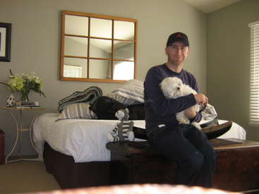 La imagen muestra a un hombre sentado en una cama, acunando a un pequeño perro blanco, con un espejo, flores y una mesa visibles en el fondo de una acogedora habitación.