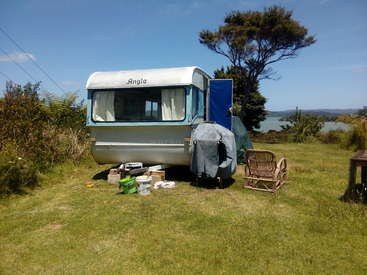La imagen muestra una caravana blanca y azul aparcada sobre hierba, con un árbol, arbustos y una masa de agua visibles al fondo bajo un cielo azul despejado.
