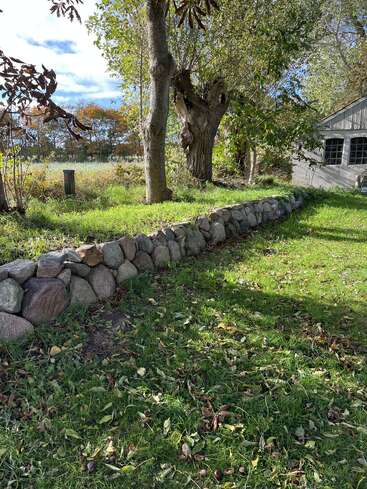 Ein sonnenbeschienener, grasbewachsener Hof mit vereinzelten Blättern, der von einer Steinmauer begrenzt wird. Ausgewachsene Bäume spenden Schatten. Ein rustikales Gebäude mit Fenstern ist teilweise auf der rechten Seite zu sehen.