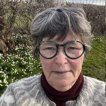 Une femme d'un certain âge, aux cheveux courts gris et portant des lunettes rondes vertes, prend un selfie en plein air. Elle porte un pull à motifs. Elle est entourée de fleurs blanches et de buissons dénudés.