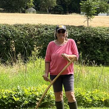 Das Bild zeigt eine ältere Frau, die in einem rosa Hemd, schwarzen Shorts und Stiefeln auf einer Wiese steht und ein langes Holzwerkzeug in der Hand hält, mit einer Hecke und Bäumen im Hintergrund.
