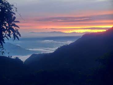 Un lever de soleil à couper le souffle sur des montagnes brumeuses, avec des teintes orange et violettes éclatantes illuminant le ciel, et une végétation luxuriante encadrant la vallée paisible et nuageuse en contrebas.