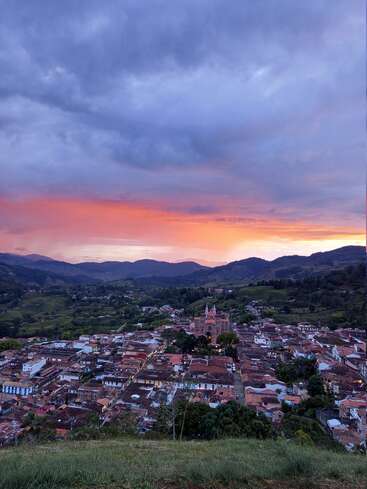Une ville animée nichée dans une vallée luxuriante au coucher du soleil, avec son église en son centre, encadrée par des nuages spectaculaires, des montagnes et un ciel orange et violet éclatant.