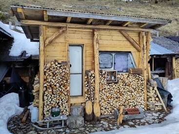 Eine kleine Holzhütte mit schrägem Dach, umgeben von ordentlich gestapeltem Brennholz. Auf dem Boden liegt Schnee, und rustikale Werkzeuge und Gegenstände schmücken die Außenfassade.