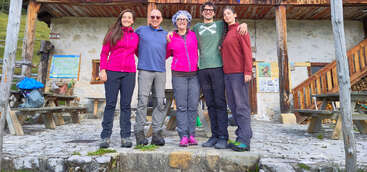Fünf Menschen stehen lächelnd vor einer rustikalen Berghütte namens „Rifugio Casera“, umgeben von Grün und Bäumen, in Wanderkleidung und genießen die Natur.