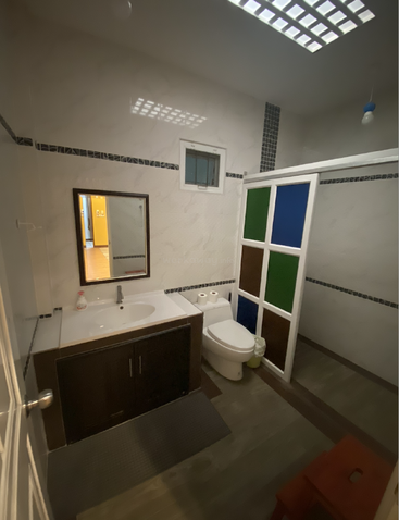Dieses Badezimmer verfügt über ein Waschbecken mit Spiegel, eine Toilette, eine bunte Trennwand, geflieste Wände und Böden, Deckenbeleuchtung, ein kleines Fenster und einen roten Hocker.
