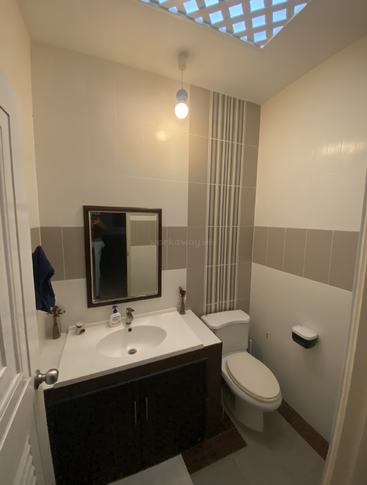 Das ist ein kleines, modernes Badezimmer mit Waschbecken, Spiegel, Toilette, weißen Fliesen, sanfter Beleuchtung und einer Decke mit einem gemusterten Fenster, durch das Tageslicht hereinfällt.
