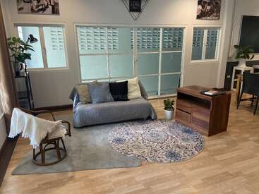 Dieses gemütliche Wohnzimmer verfügt über ein graues Sofa mit Kissen, einen Holzstuhl, einen runden gemusterten Teppich, Pflanzen, große Fenster und eine minimalistische Einrichtung mit Wandbildern.