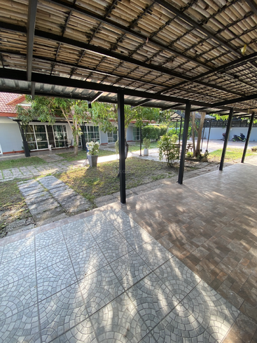 Eine überdachte Terrasse mit gemusterten Fliesen bietet einen Blick auf einen Gartenbereich mit Topfpflanzen, Bäumen und einem Weg, der zu einem modernen Haus mit großen Fenstern führt.