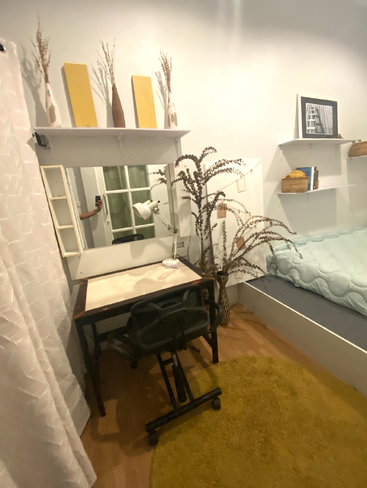 Ein gemütliches, minimalistisch eingerichtetes Schlafzimmer verfügt über einen Schreibtisch mit Spiegel, einen Drehstuhl, gelbe Akzente, Trockenpflanzen, weiche Bettwäsche, Regale und einen runden, senffarbenen Teppich.