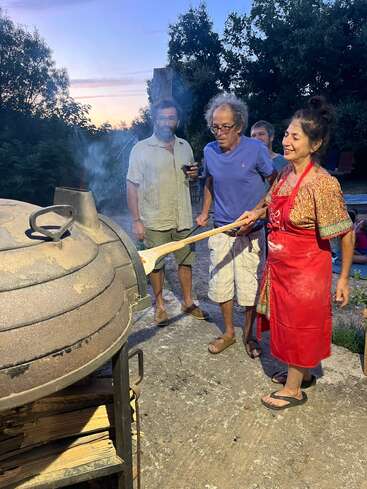 Quatre adultes se rassemblent à la tombée de la nuit autour d'un four à bois rustique. Une femme, souriante, vêtue d'un tablier rouge, utilise une pelle en bois pour cuire quelque chose.