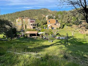 Des maisons colorées se nichent dans une vallée verdoyante et luxuriante. Les gens se rassemblent sous un abri et des parasols, profitant d'une journée ensoleillée, entourés d'arbres et de collines pittoresques.