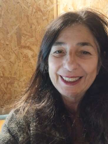 Une femme aux longs cheveux bruns sourit à la caméra. Elle se trouve à l'intérieur, debout devant un mur en bois texturé, l'air heureux et détendu.