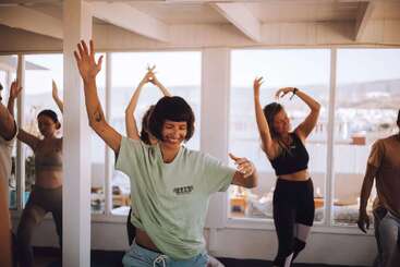 A group of people joyfully dance together indoors, surrounded by large windows with scenic views. Everyone looks happy, engaged, and connected, embracing a lively, communal atmosphere.