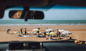 This photo, taken from inside a car, shows people enjoying a sunny day on a sandy beach with umbrellas, towels, surfboards, and the ocean in the background.