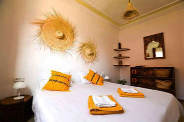 A cozy bedroom with a white bed, yellow pillows and towels, straw hats as wall decor, rustic wooden furniture, woven baskets, shelves, and a decorative mirror.