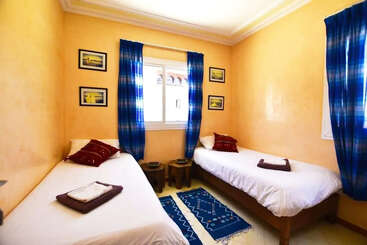 This cozy bedroom features two single beds with white linens, colorful pillows, blue curtains, yellow walls, decorative wall art, a window, and a vibrant blue rug.