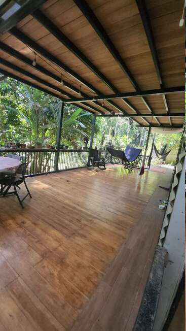 This image shows a spacious wooden porch with hammocks, chairs, a table, potted plants, and lush green trees outside, creating a relaxing, inviting tropical atmosphere.