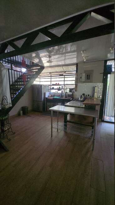 This image shows a modern kitchen with wooden floors, stainless steel countertops, appliances, large windows, a staircase, and a ceiling fan, creating an airy, open atmosphere.