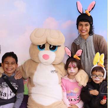 Un grupo de niños sonrientes y un adulto, con orejas de conejo, posan con un gran y simpático conejo mascota. El ambiente es festivo y alegre, probablemente de Pascua.