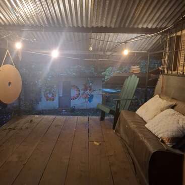 Um pátio aconchegante e rústico com piso de madeira, teto de metal corrugado, luzes de corda, uma cadeira verde, um sofá almofadado e arte colorida na parede, criando um ambiente acolhedor.