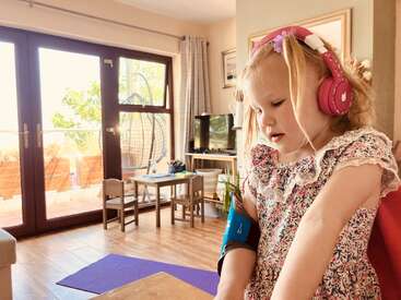 Uma menina com fones de ouvido cor-de-rosa e um vestido florido está sentada dentro de casa. Ela parece concentrada, talvez brincando ou estudando, em uma sala de estar bem iluminada, aconchegante e ensolarada.