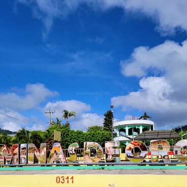 Grandes letras coloridas escrevem "MINAS DE ORO", decoradas com imagens locais. Palmeiras, vegetação exuberante, linhas de energia e um prédio branco estão sob um céu brilhante e vibrante.