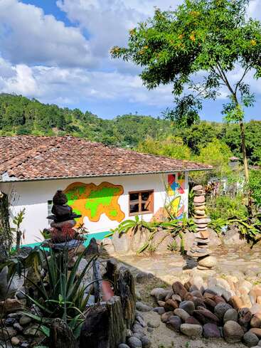 Una colorida casa rural con tejado de tejas se asienta en medio de una exuberante vegetación. En el jardín hay piedras apiladas, plantas y un mural con un mapamundi.