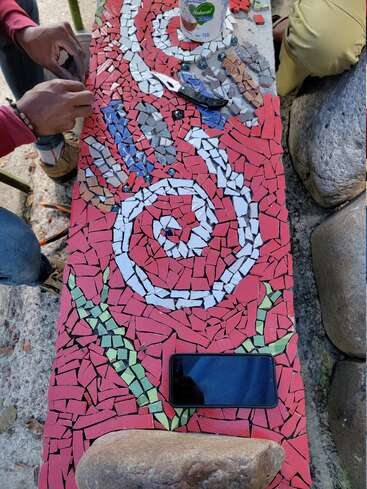 Um mosaico colorido está sendo montado em um banco com azulejos vermelhos, brancos, verdes, azuis e cinza. Mãos, celular, ferramentas, pedras e recipiente visíveis.