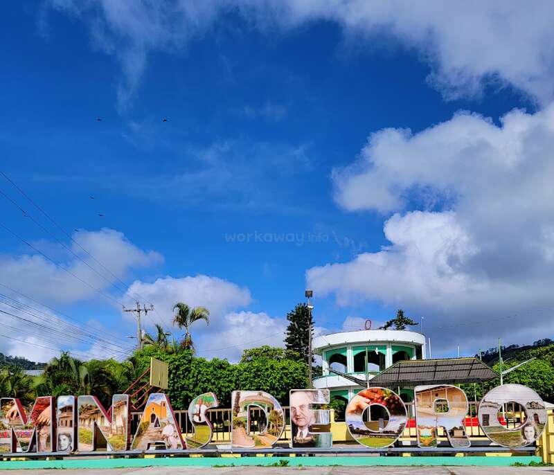 Large colorful letters spell “MINAS DE ORO,” decorated with local images. Palm trees, lush greenery, power lines, and a white building stand beneath a bright, vibrant sky.