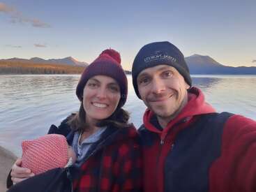 A smiling couple and a baby pose for a selfie by a serene lake, with mountains in the background, all wearing cozy hats and warm clothes.