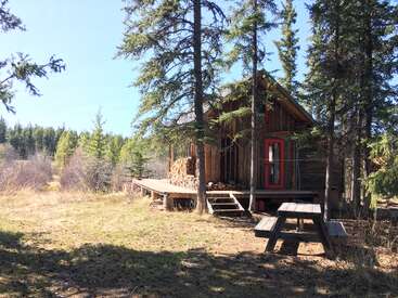 A rustic wooden cabin sits among tall pine trees, with stacked firewood on the porch and a picnic table nearby, surrounded by nature on a sunny day.
