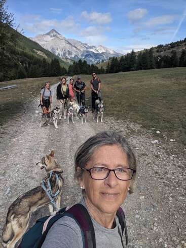 A woman takes a selfie while hiking with a group of people and several huskies. Majestic mountains and green forests form a scenic background.