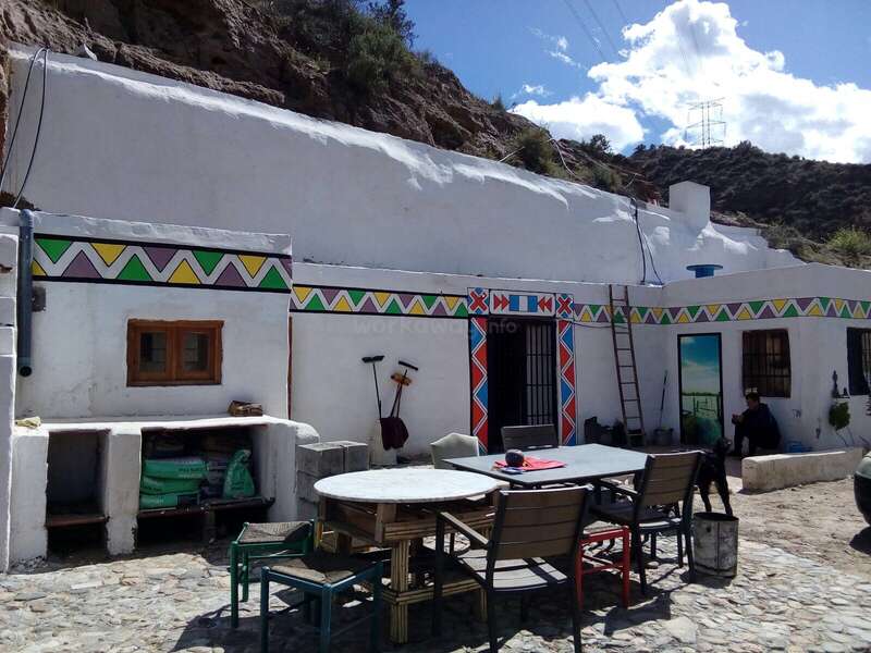 A imagem mostra um edifício branco com padrões geométricos coloridos no exterior, com um pátio com mesa e cadeiras, cercado por uma encosta rochosa sob um céu azul com nuvens.