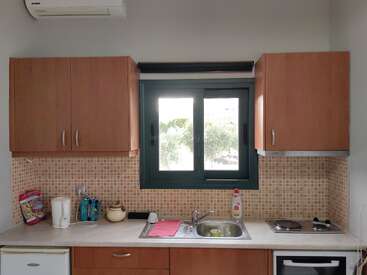 Das Bild zeigt eine kleine Küche mit Holzschränken, einem Fenster in der Mitte, einer Spüle, einem Herd, einem Wasserkocher, Spülmittel und einer gefliesten Rückwand. Sauber und aufgeräumt.