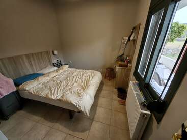 Ein kleines, aufgeräumtes Schlafzimmer mit einem Doppelbett, beigen Wänden, Fliesenboden, einem Fenster, einem Heizkörper, einem Schminktisch mit Spiegel, Koffern und einigen persönlichen Gegenständen, die herumliegen.
