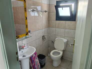 Das ist ein kleines, gefliestes Badezimmer mit einer weißen Toilette, einem offenen Fenster, einem Waschbecken, Seife, einem Spiegel mit Sonnen- und Mondmotiv, Toilettenpapier und einem Mülleimer.