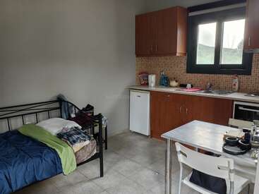 Ein kleines Studio-Apartment verfügt über ein Einzelbett, eine einfache Küchenzeile mit Holzschränken, einen Tisch mit Stühlen, einen Blick aus dem Fenster und hier und da verstreute Dinge des täglichen Bedarfs.