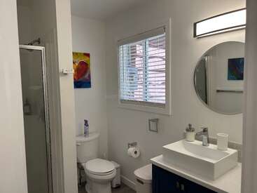 Dieses Bild zeigt ein sauberes, modernes Badezimmer mit einer Glasdusche, einer weißen Toilette, einem modernen Waschbecken, einem runden Spiegel, einem Fenster mit Jalousien und bunten Kunstwerken an der Wand.