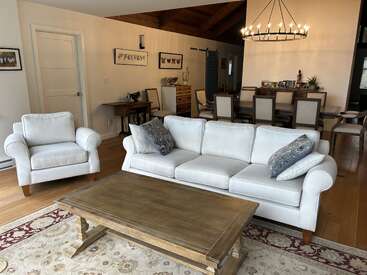 Dieses gemütliche Wohnzimmer verfügt über ein weißes Sofa, einen Sessel, einen Couchtisch aus Holz, einen Essbereich, einen Kronleuchter, einen gemusterten Teppich, Wandkunst und einen warmen Holzfußboden. Einladendes Ambiente.