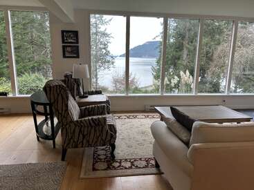 Ein gemütliches Wohnzimmer mit großen Fenstern, gemusterten Stühlen und einem Blick auf Bäume und Wasser. Die Natur umgibt den Raum und schafft eine friedliche, einladende Atmosphäre zum Entspannen.